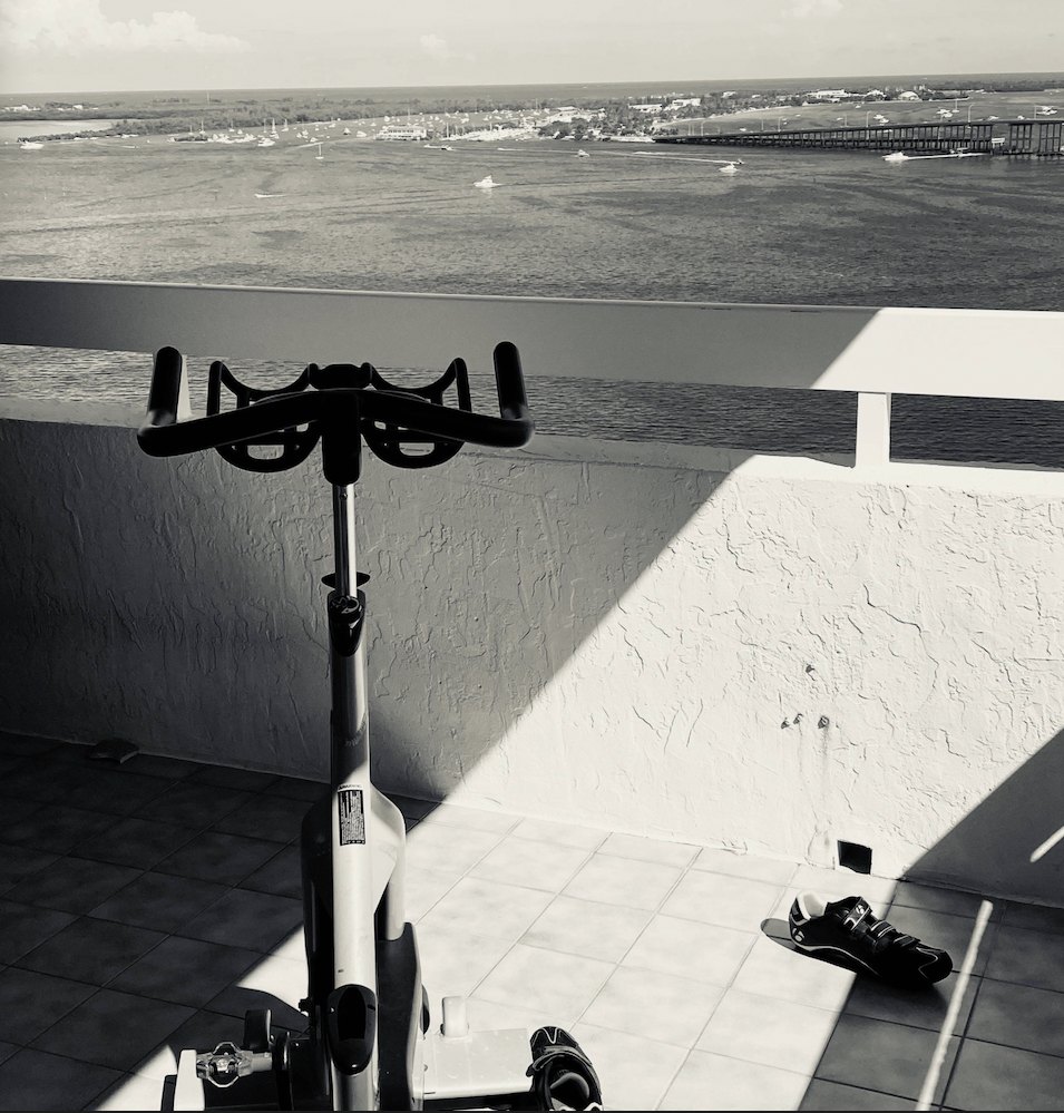 Black-and-white photograph: an exercise bike on a sunlit balcony overlooking a bay with boats and a bridge